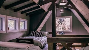 Una camera da letto con soffitto spiovente, un letto con biancheria a motivi, due finestre e vista su alberi.