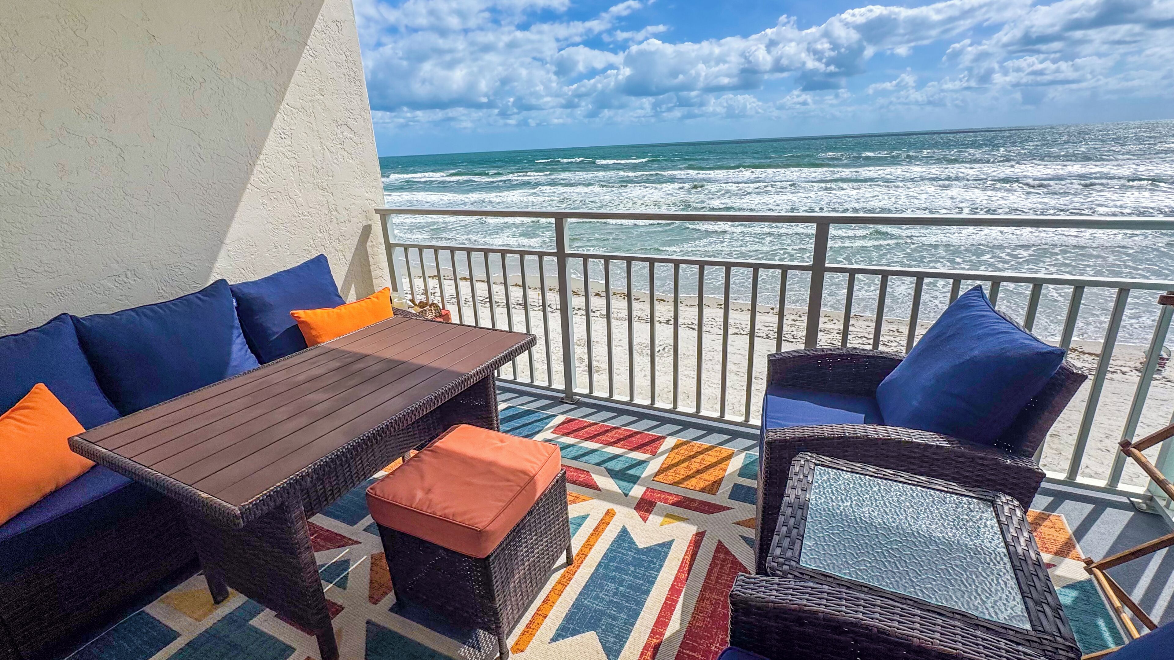 A balcony with wicker furniture, a wooden table, and a colorful rug.