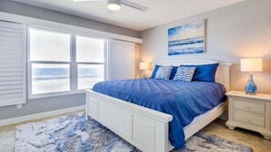 A bedroom with a large window, a bed with blue bedding, a nightstand, and a lamp.