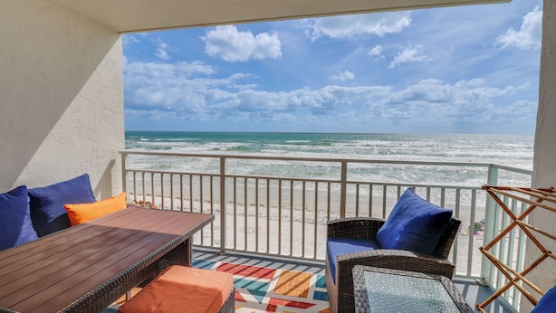 A balcony with wicker furniture, a wooden table, and colorful cushions, overlooking a beach with waves and a clear sky.