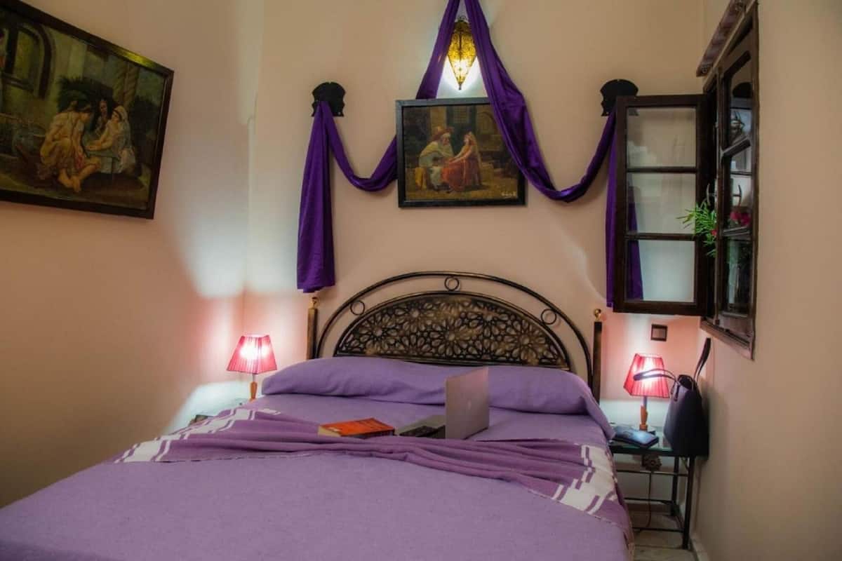 A bedroom with a bed, a laptop, two bedside lamps, a painting, and a window with curtains.