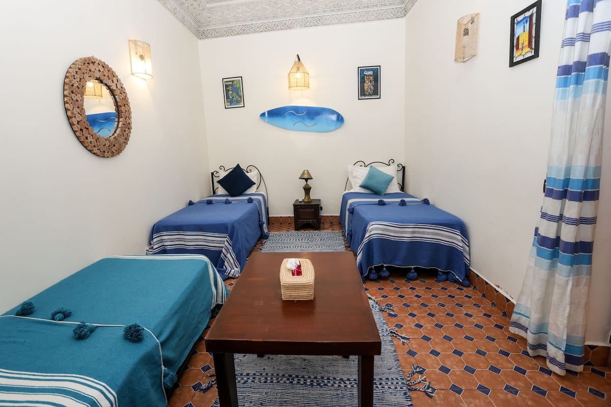 A room with two single beds, a small table, a wall-mounted mirror, and a decorative wall hanging.