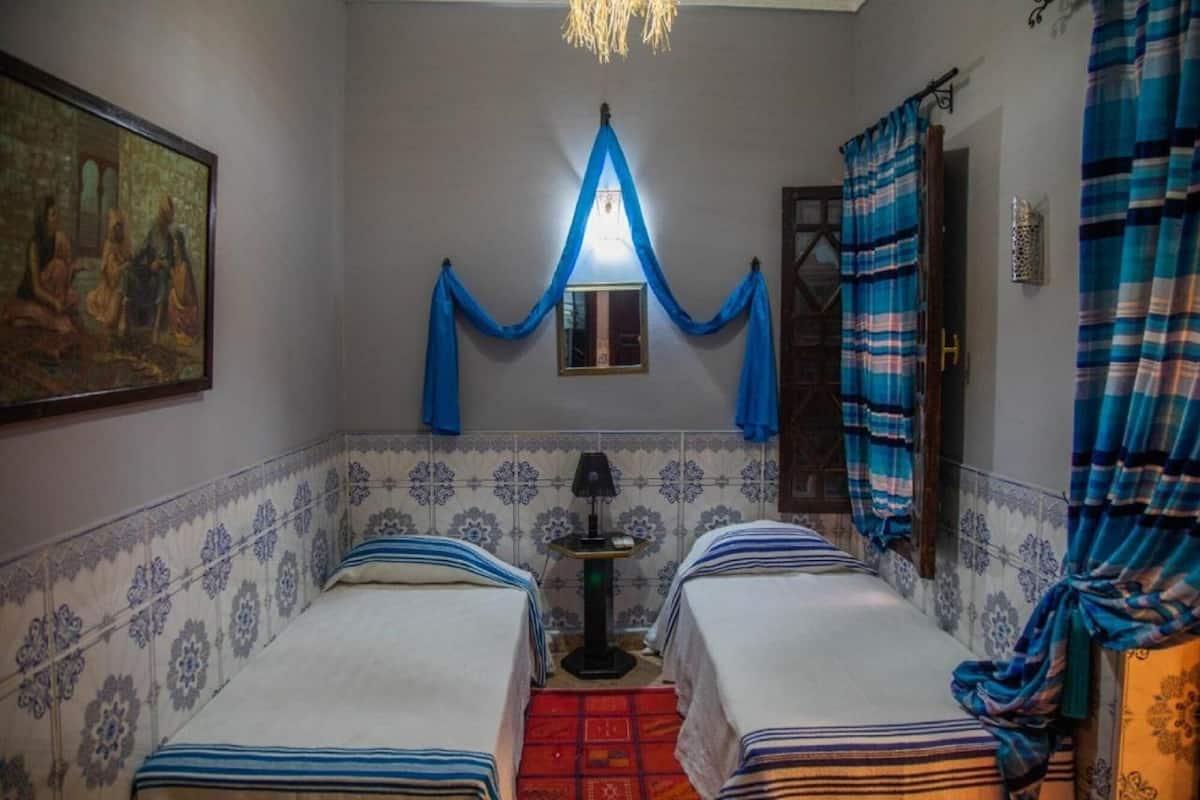 A room with two beds, a small table, a lamp, and a painting on the wall.