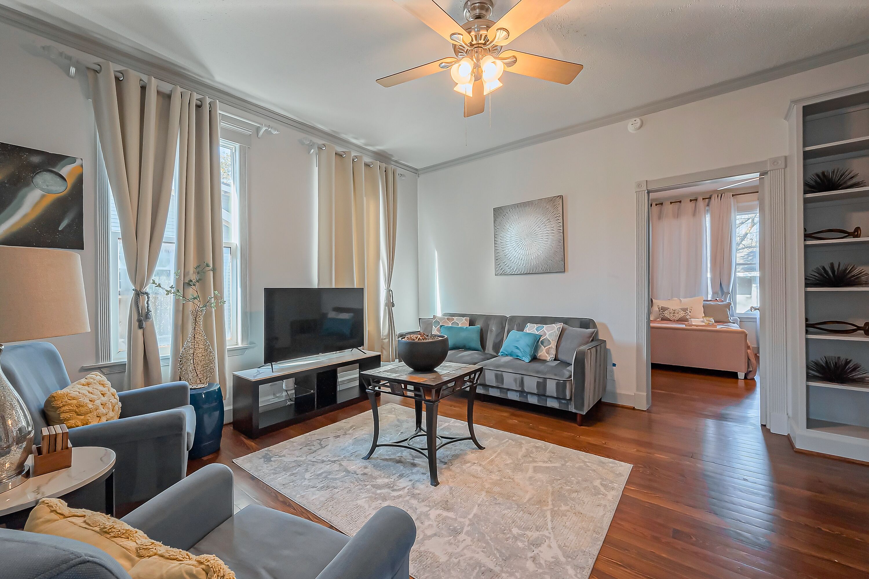 A living room with a TV, sofa, and chairs, a ceiling fan, and a bookshelf.