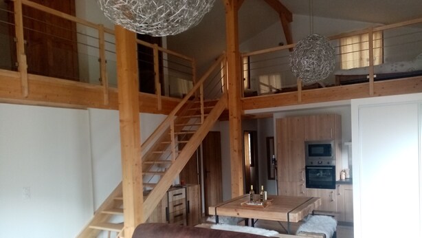 A spacious living area with a brown leather sofa, wooden staircase, and a kitchen area with a dining table.