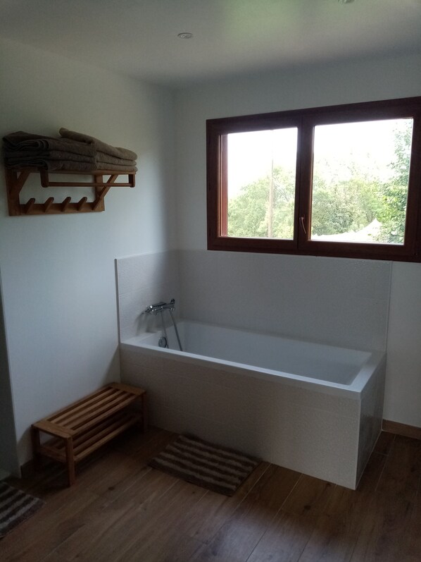 A bathroom with a bathtub, wooden bench, and a window with a view of greenery.
