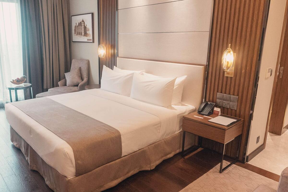 A hotel room with a large bed, a bedside table with a phone and a notepad, a chair, a small table with a bowl, and a window with curtains.