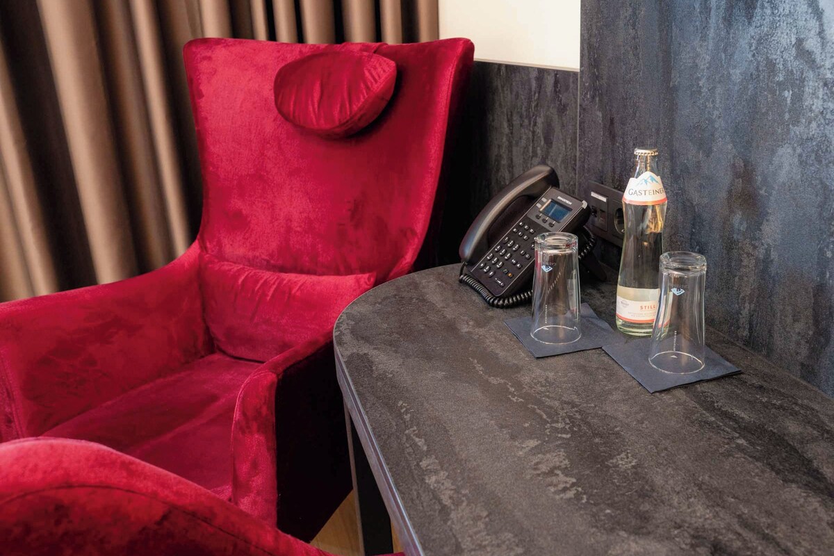 A red velvet chair with a pillow, a black telephone, and two glass bottles on a dark table.