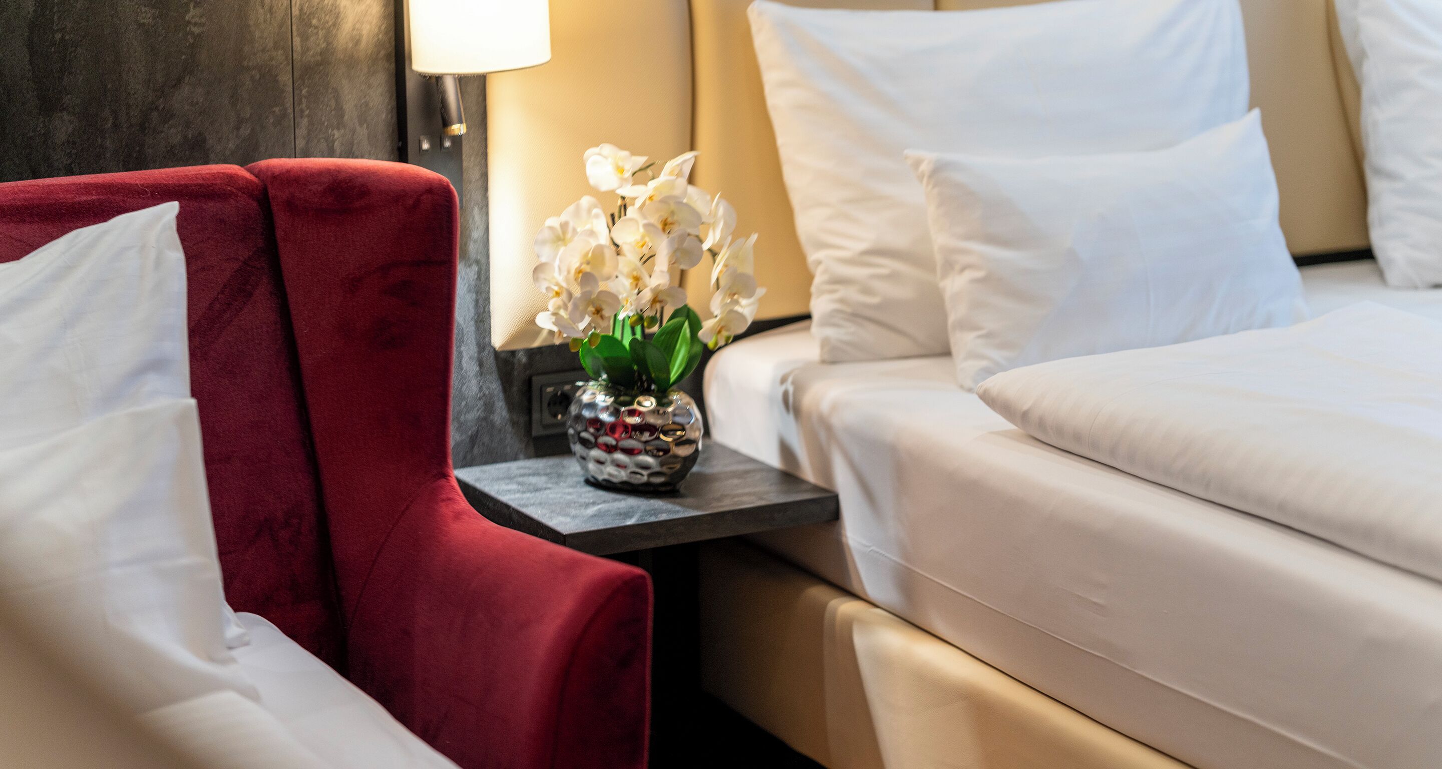 Quarto de hotel com poltrona vermelha, uma mesa pequena com um vaso decorativo e uma cama com roupa de cama branca.