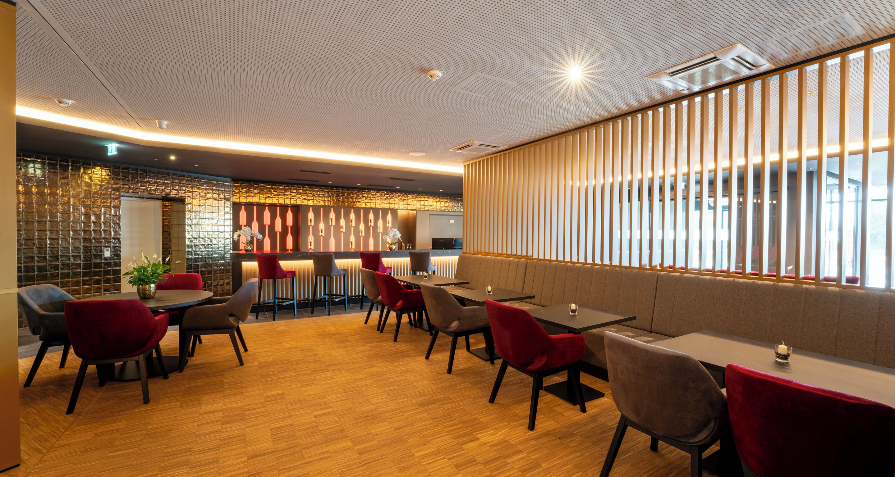 Un restaurante moderno con suelo de madera, sillas rojas y grises, y una zona de bar con una pared decorativa.