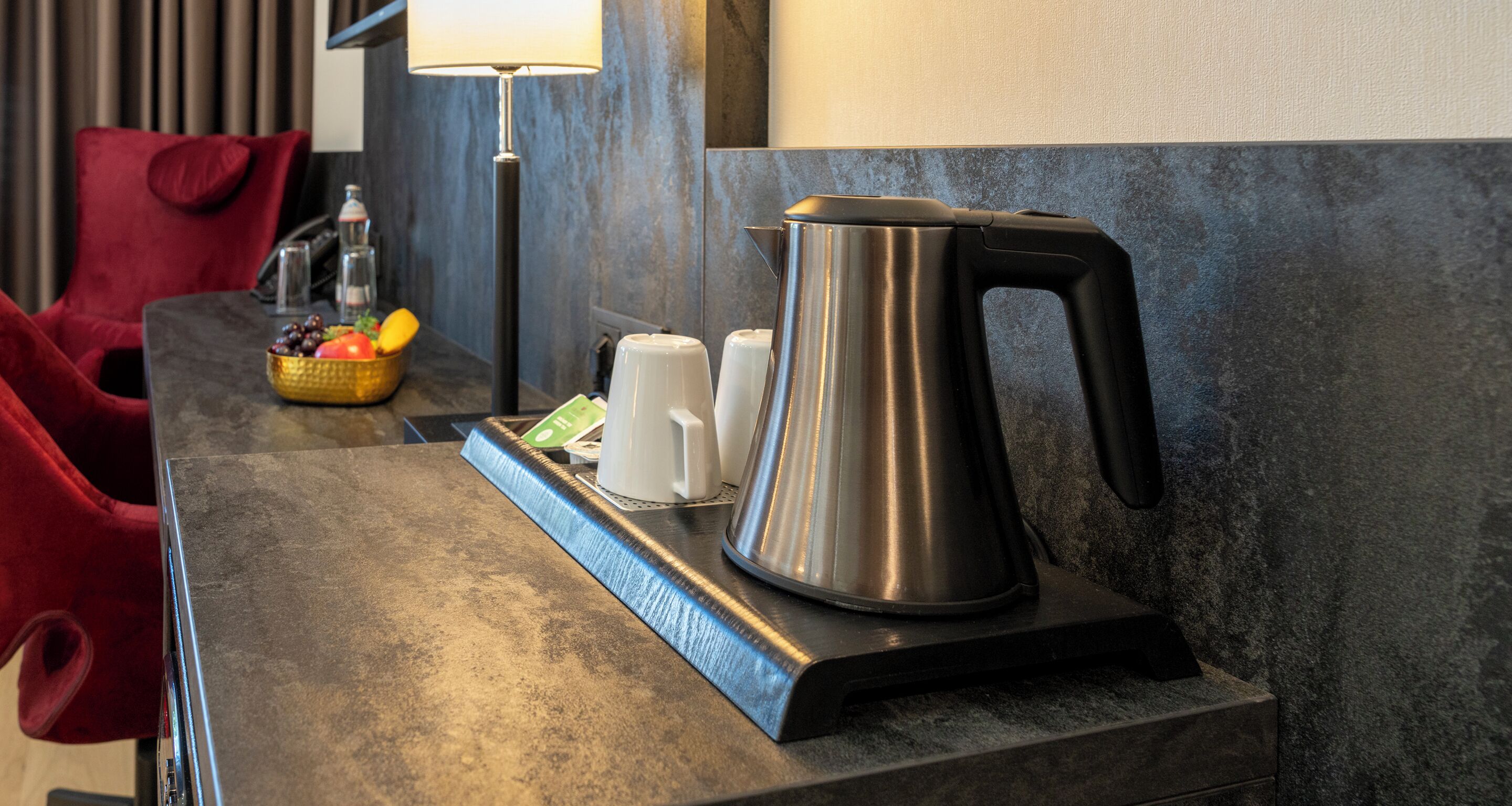 A modern hotel room with a metallic kettle, white cups, and a fruit basket on a table.