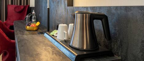 A modern hotel room with a metallic kettle, white cups, and a fruit basket on a table.