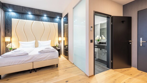 A modern hotel room with a large bed, bedside lamps, a dark door, and a view into the bathroom. - PLAZA INN Wiener Neustadt, BW Signature Collection (Wiener Neustadt)