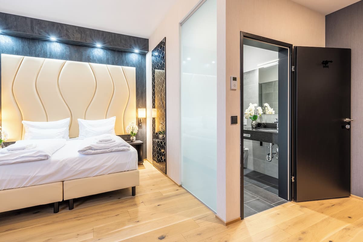 Ein modernes Hotelzimmer mit einem großen Bett, Nachttischlampen, einer dunklen Tür und Blick ins Badezimmer.