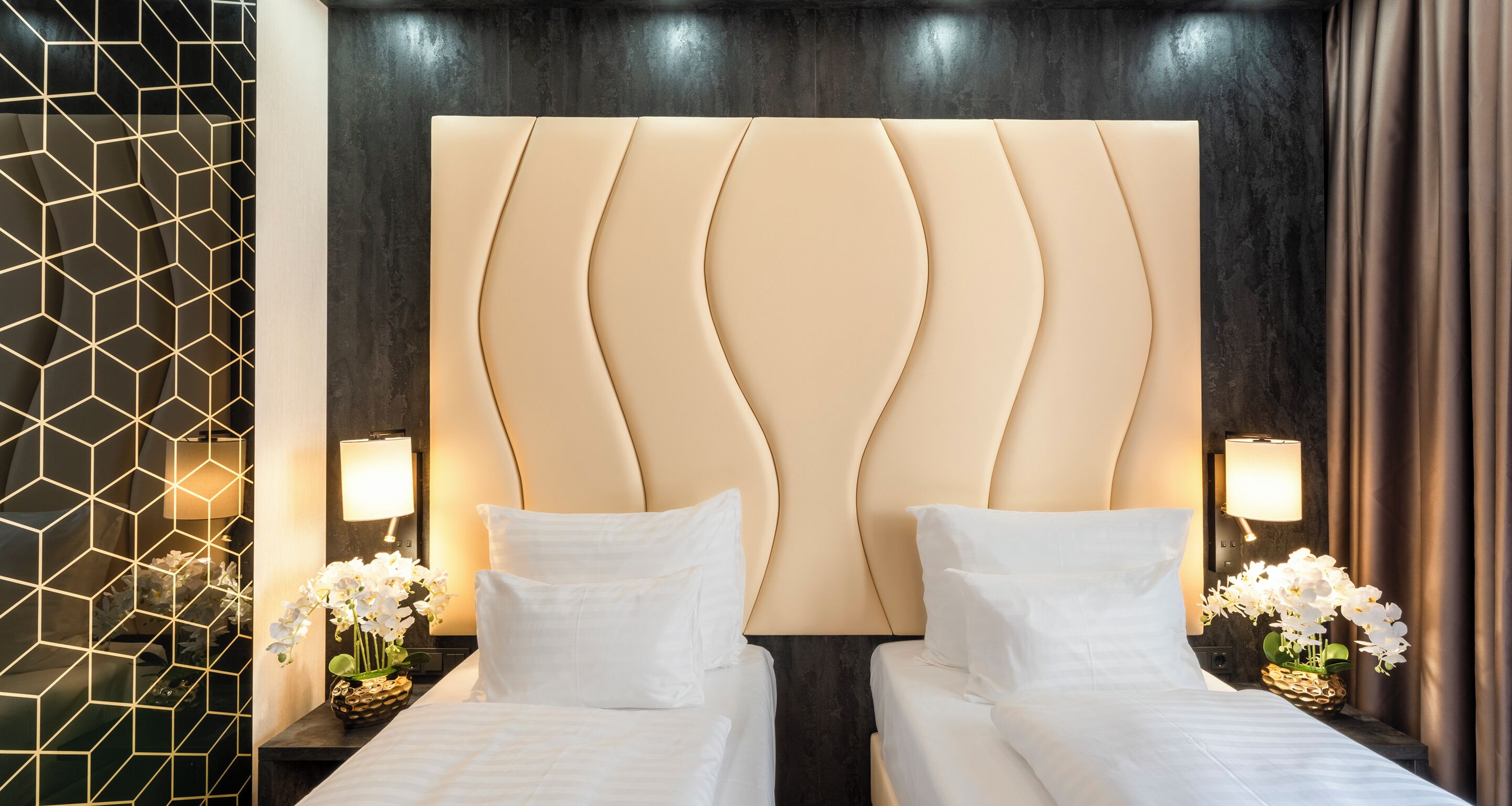A hotel room with a large, cushioned headboard, two beds with white linens, bedside lamps, and a decorative wall panel.
