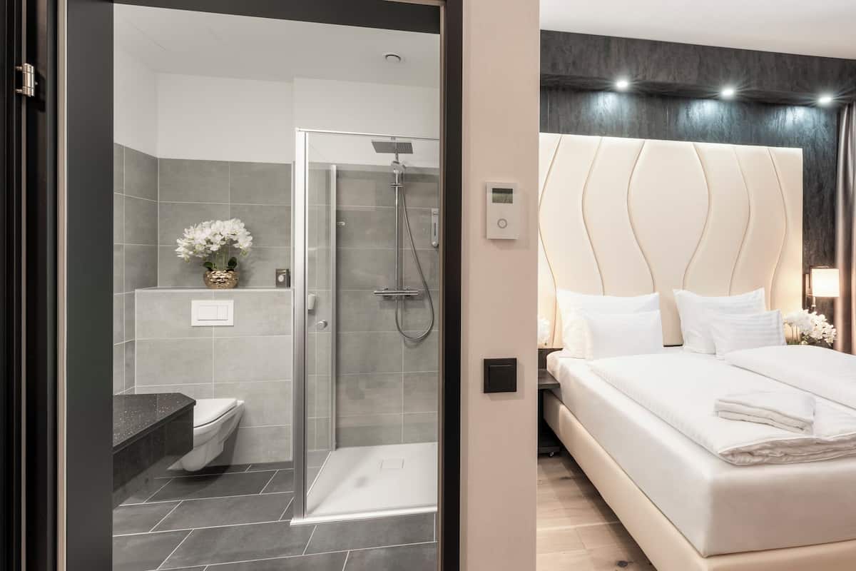 Ein modernes Hotelzimmer mit einer gläsernen Duschkabine, einem großen Bett mit weißen Laken und einem dunklen Kopfteil.