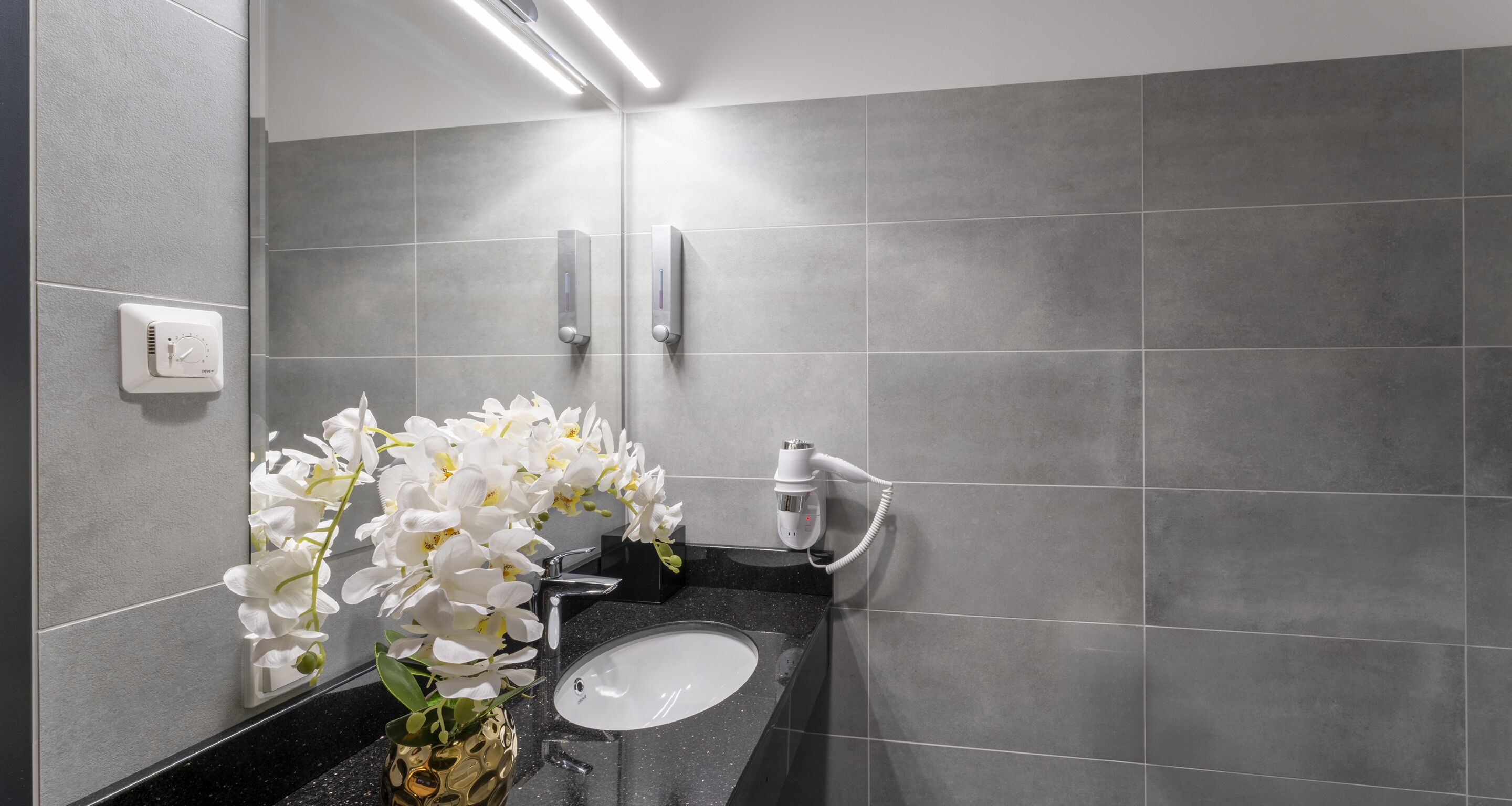 Casa de banho moderna com bancada preta, lavatório branco e espelho. Há dois dispensadores de toalhas fixados na parede e um secador de cabelo.