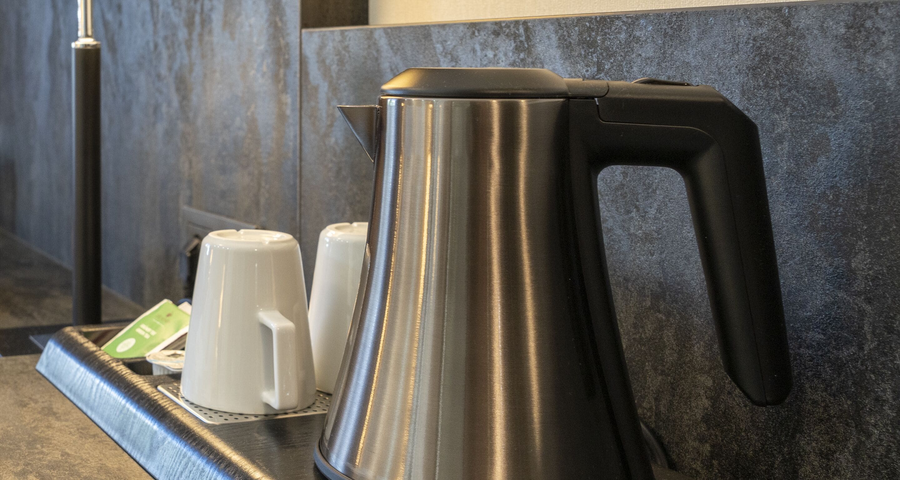 A stainless steel kettle on a tray with two white cups and a green card, set against a textured wall.