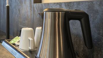 A stainless steel kettle on a tray with two white cups and a green card, set against a textured wall.