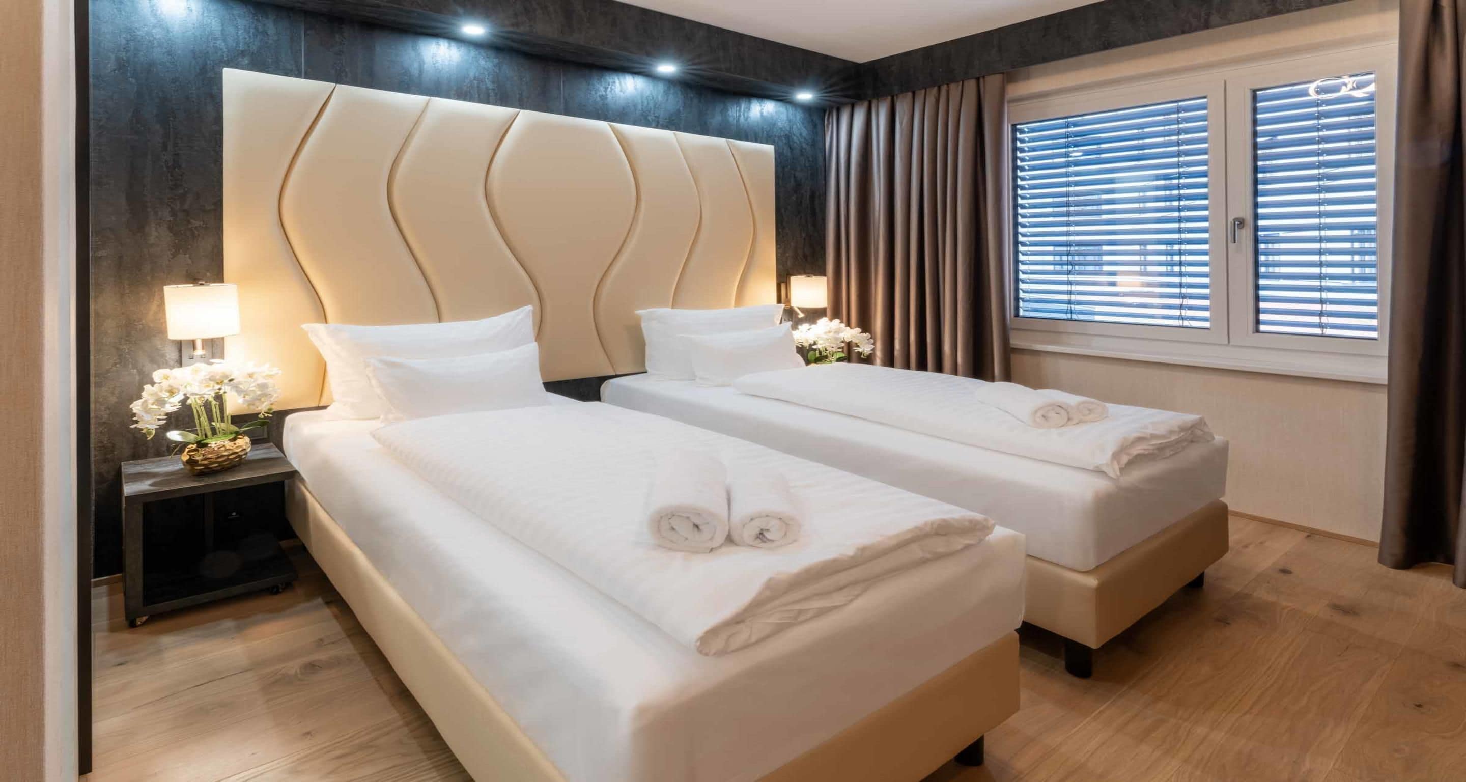 Een hotelkamer met twee bedden, een groot hoofdeinde, bedlampen en een raam met zonwering.