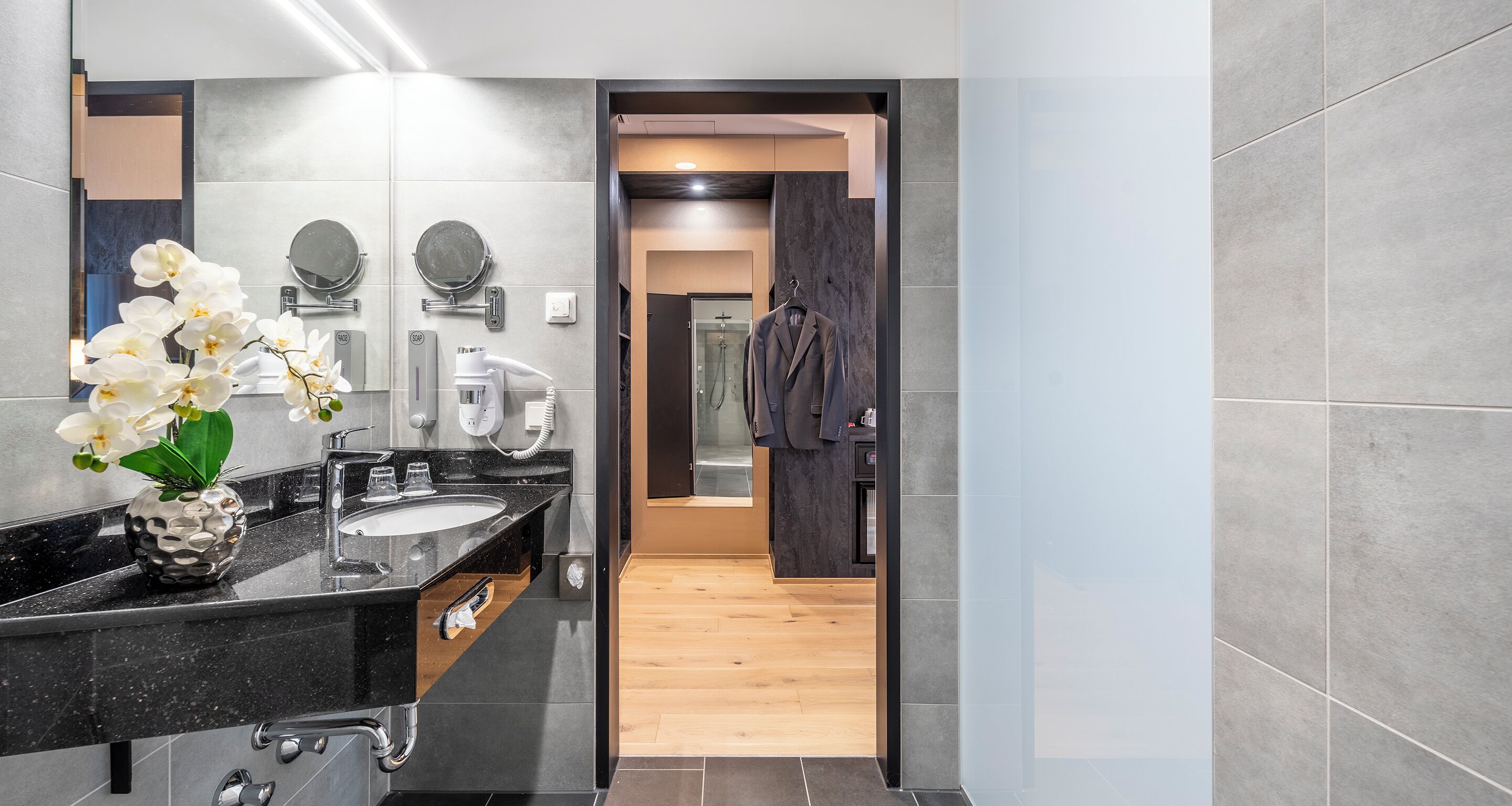 Un baño moderno con encimera negra, un espejo redondo, toallero y zona de ducha visible a través de una puerta abierta.