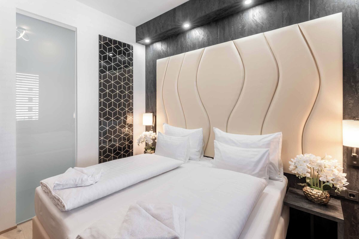 A hotel room with a large bed, a bedside table with a lamp, a decorative wall panel, and a door with frosted glass.