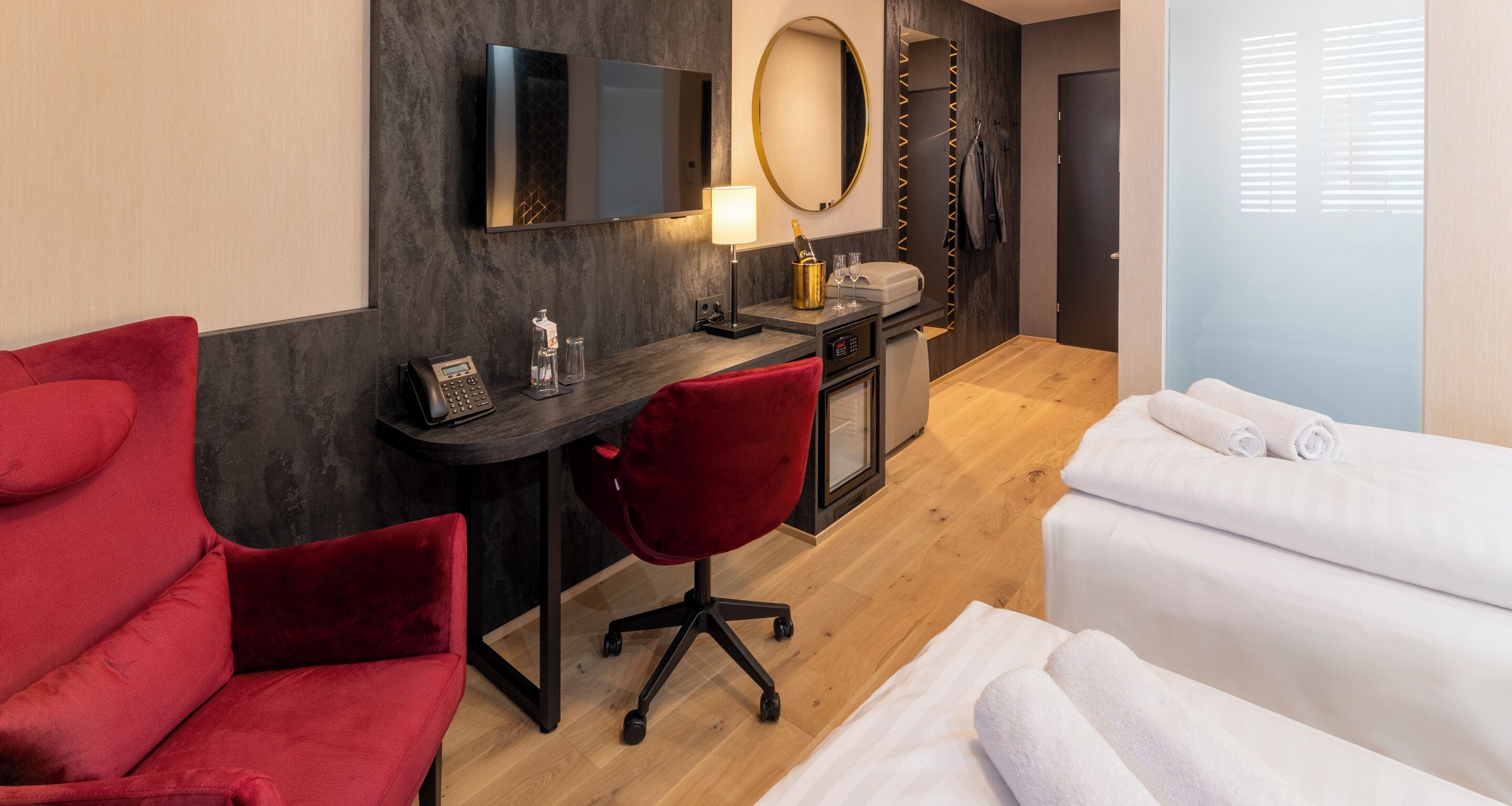 A hotel room with a desk, a red chair, a bed with white linens, a TV, a mirror, and a minibar.