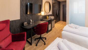 A hotel room with a desk, a red chair, a bed with white linens, a TV, a mirror, and a minibar.