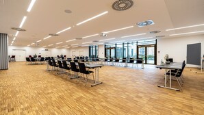 A spacious conference room with a wooden floor, multiple tables with chairs, and large windows offering a view of the outdoors. - PLAZA INN Wiener Neustadt, BW Signature Collection (Wiener Neustadt)