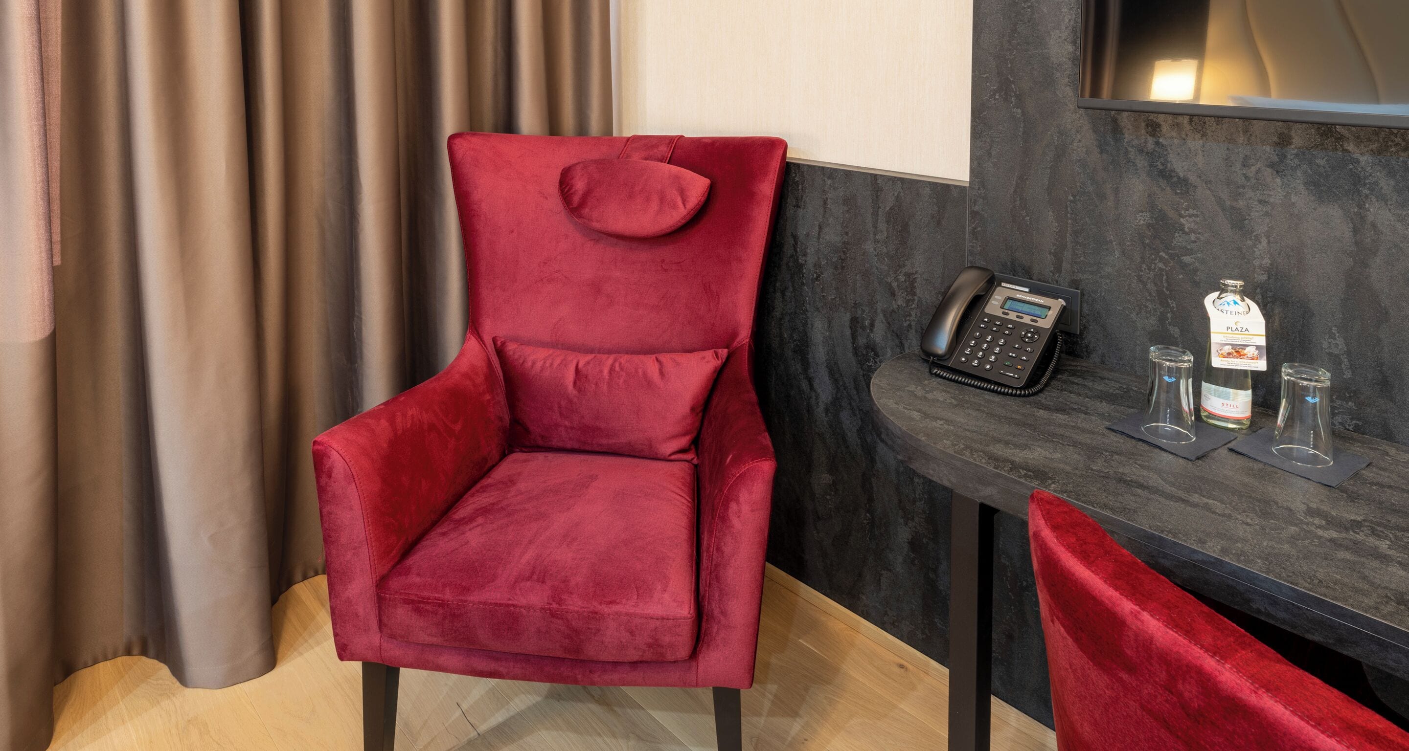 Ein roter Samtstuhl mit gepolstertem Sitz und Rückenlehne, ein schwarzes Telefon und zwei Gläser mit einer Flasche Stein Plaza Wein auf einem dunklen Marmor-Tisch.
