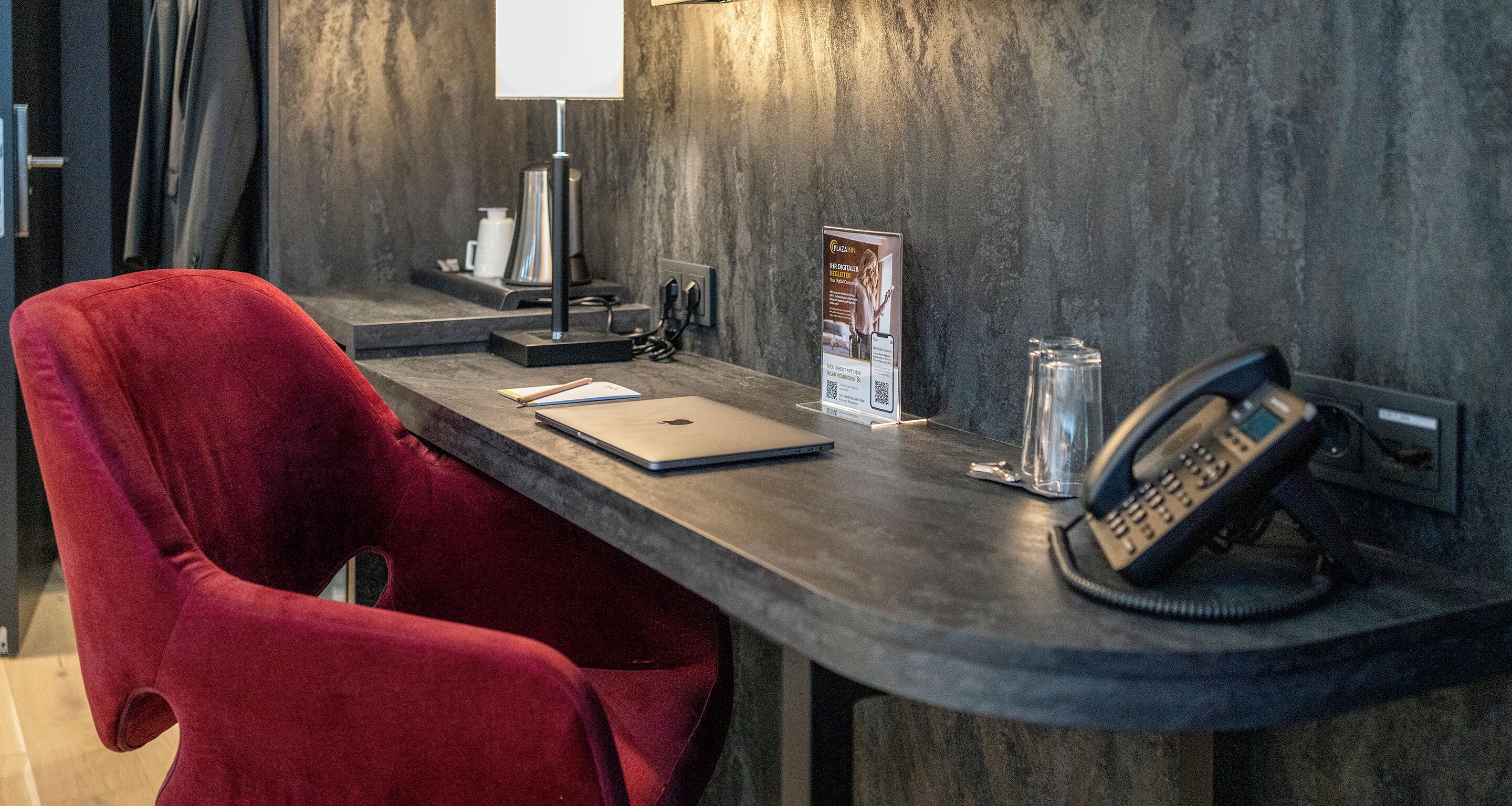 Ein Hotelrezeptionstresen mit einem roten Sessel, einem Laptop, einem Telefon, einer Lampe und einer Broschüre.