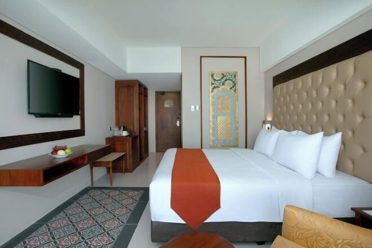 A hotel room with a large bed, a flat-screen TV mounted on the wall, a wooden desk with a fruit bowl, a patterned rug, and a decorative door.