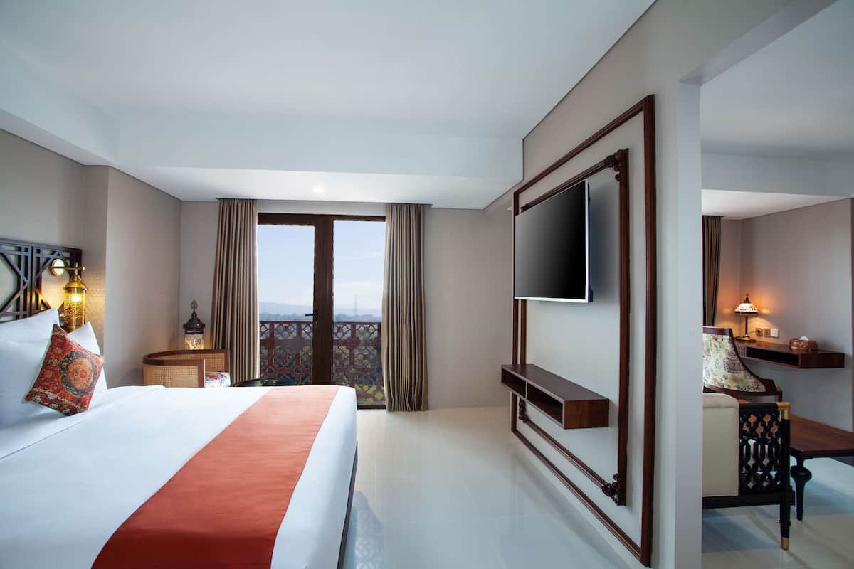 Une chambre d’hôtel avec un grand lit, un téléviseur fixé au mur, un bureau avec une chaise et un balcon avec des rideaux.
