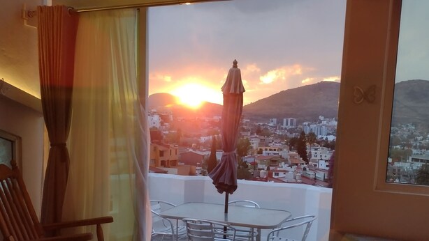 Un balcón con una mesa y sillas, una mecedora de madera, y vistas a un paisaje urbano al atardecer.