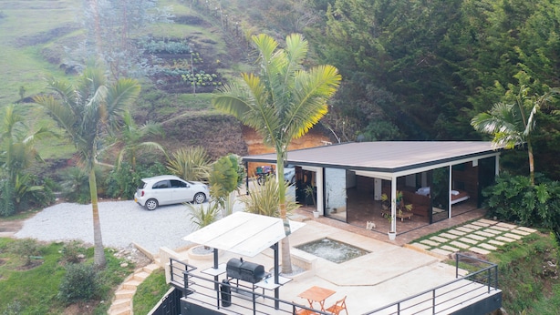A modern house with a covered patio, a car parked in front, and a lush green landscape in the background.