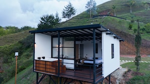 A modern, elevated cabin with a wooden deck, large windows, and a view of a hilly landscape.