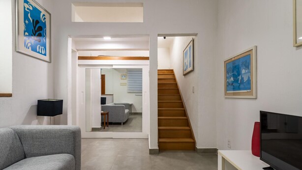 Una sala moderna con un sofá gris, un televisor sobre un mueble bajo y una escalera que conduce a otra habitación.