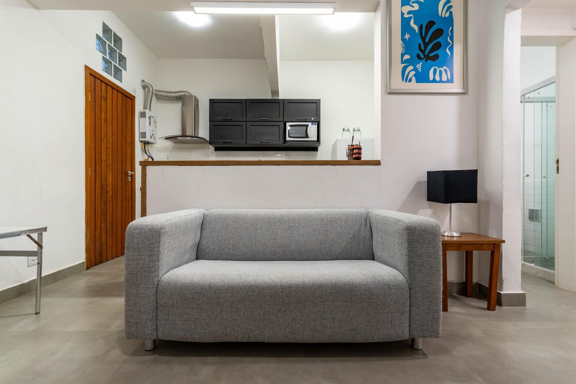 Un salon moderne avec un canapé gris, une petite table en bois surmontée d’une lampe, un coin cuisine doté de meubles noirs et d’un four à micro-ondes, et une œuvre d’art encadrée accrochée au mur.