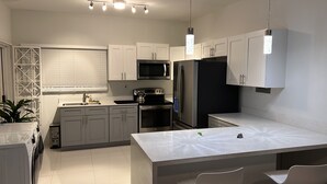 Cocina moderna con armarios blancos, un refrigerador de acero inoxidable, un microondas y un horno empotrado. Hay una isla grande con un fregadero y una tetera en la estufa.