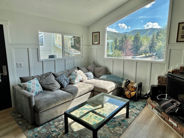 A living room with a sofa, a glass coffee table, and a large window offering a mountain view.