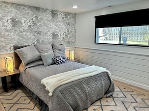 A bedroom with a gray quilted bedspread, a white throw blanket, and a cloud-patterned wall. There are two bedside tables with lamps, a window with a black blind, and a patterned rug on the floor.
