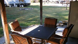 An outdoor patio with a table and four chairs, a refrigerator, and a grassy area with a parked RV in the background.