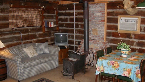 A cozy log cabin interior with a checkered sofa, a small table with a floral tablecloth, a fireplace, and a television.