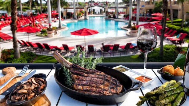 Una mesa de comedor con un bistec a la parrilla, espárragos y una copa de vino tinto, con vista a un área de piscina con sombrillas y sillones rojas.