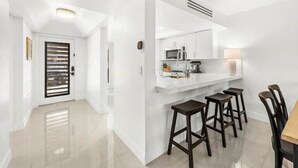 Cocina moderna con gabinetes blancos, microondas y cafetera. Hay cuatro taburetes y una mesa con sillas.