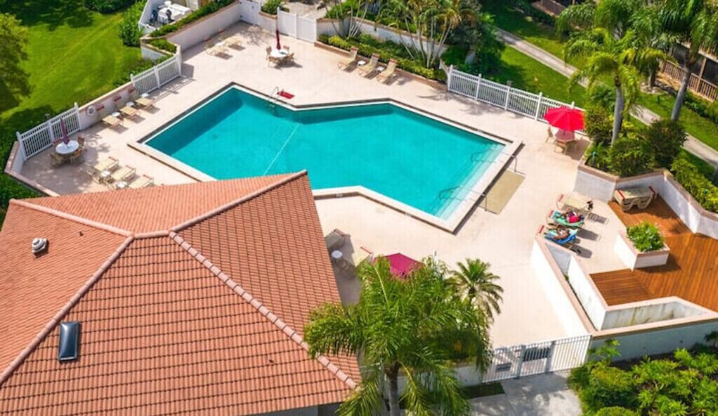 A rooftop swimming pool with a tiled roof, surrounded by a white fence, and equipped with sun loungers and umbrellas.