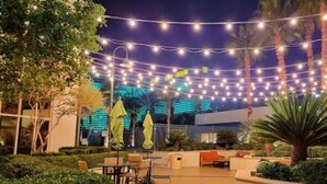 An outdoor seating area with string lights, palm trees, and a building with illuminated windows in the background.