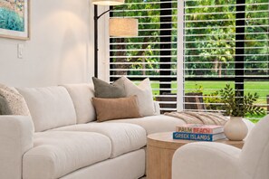 A modern living room with a white sofa, a round wooden side table, and a book titled 'Palm Beach Greek Islands'.