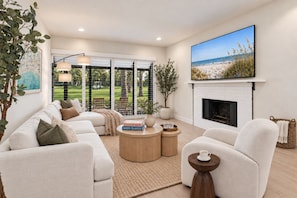 A spacious living room with a white fireplace, a large flat-screen TV, and a view of a golf course through large windows. - New Home at PGA National on Golf Course with Private Spa by Guaranteed Rental™ (Palm Beach Gardens)