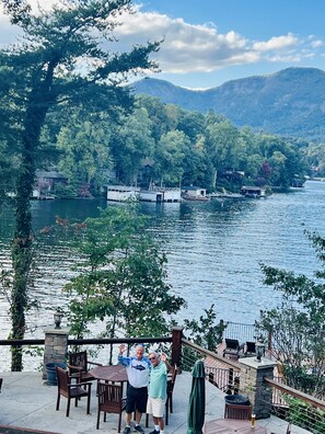 A lakeside patio with wooden tables and chairs, overlooking a calm lake and forested mountains.